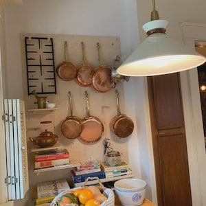 Op de afbeelding: Een lichtgekleurde houten pegboard wanddisplay met diverse koperen steelpannen met messing handgrepen, een zwart metalen fornuisrooster, een koperen ketel, gestapelde kookboeken, een keramische vijzel met stamper en een witte hanglamp.