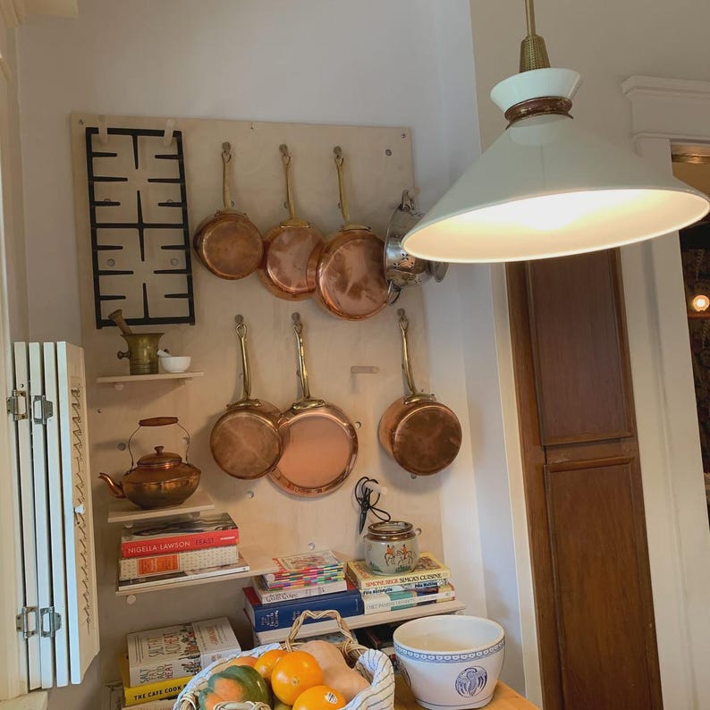 Op de afbeelding: Een lichtgekleurde houten pegboard wanddisplay met diverse koperen steelpannen met messing handgrepen, een zwart metalen fornuisrooster, een koperen ketel, gestapelde kookboeken, een keramische vijzel met stamper en een witte hanglamp.