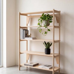 May include: A light-colored wooden bookshelf with four shelves featuring vertical side supports. The shelves hold a trailing green plant in a gray pot, a stack of books with a gold-toned dog figurine, a snake plant in a black pot, and two books.