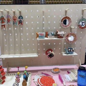 A light wood pegboard display stand with various colorful ceramic items, including mugs, plates, and figurines, displayed on shelves and pegs.