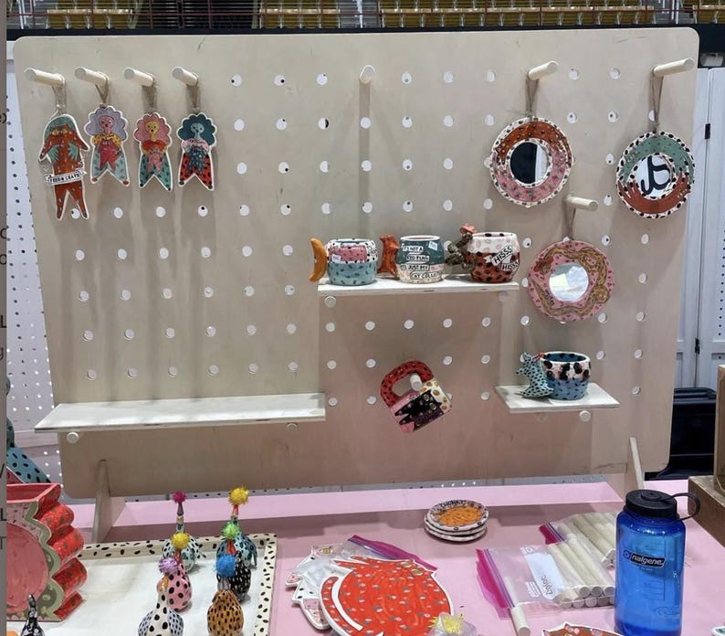 A light wood pegboard display stand with various colorful ceramic items, including mugs, plates, and figurines, displayed on shelves and pegs.