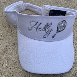 May include: White visor with a rhinestone design of a tennis racket and the word "Holly".