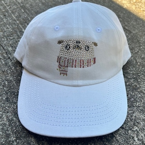 May include: White baseball cap with a decorative rhinestone design of a dog wearing a scarf. The dog design is made of silver, black, red, and gold rhinestones. The cap has a curved brim and a button on top.