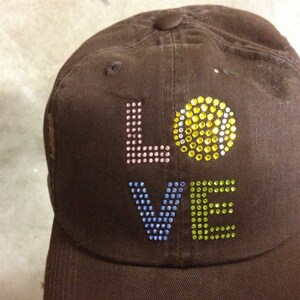 May include: A brown baseball cap with the word "LOVE" spelled out in colorful rhinestones. The "O" is a large yellow rhinestone.