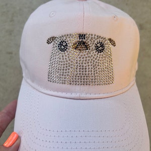 May include: A light pink baseball cap with a rhinestone design of a pug's face. The pug's face is made of silver rhinestones, with black rhinestones for the eyes and nose. The cap has a curved brim and a button on top.