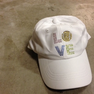 May include: White baseball cap with the word "LOVE" spelled out in colorful rhinestones. The "O" is replaced with a yellow tennis ball.