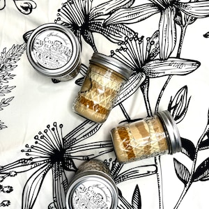 May include: Three glass jars with silver lids containing a layered mixture of brown and white ingredients. The jars are labeled "Remarkable Complete Wellness" and are arranged on a white background with black floral designs.