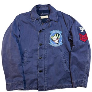 Op de afbeelding: Marineblauwe vintage jas met knoopsluiting, kraag en twee voorzakken. De jas heeft patches met tekst en een ram-afbeelding, samen met een militair insigne op de mouw. Gemaakt van een duurzame stof.