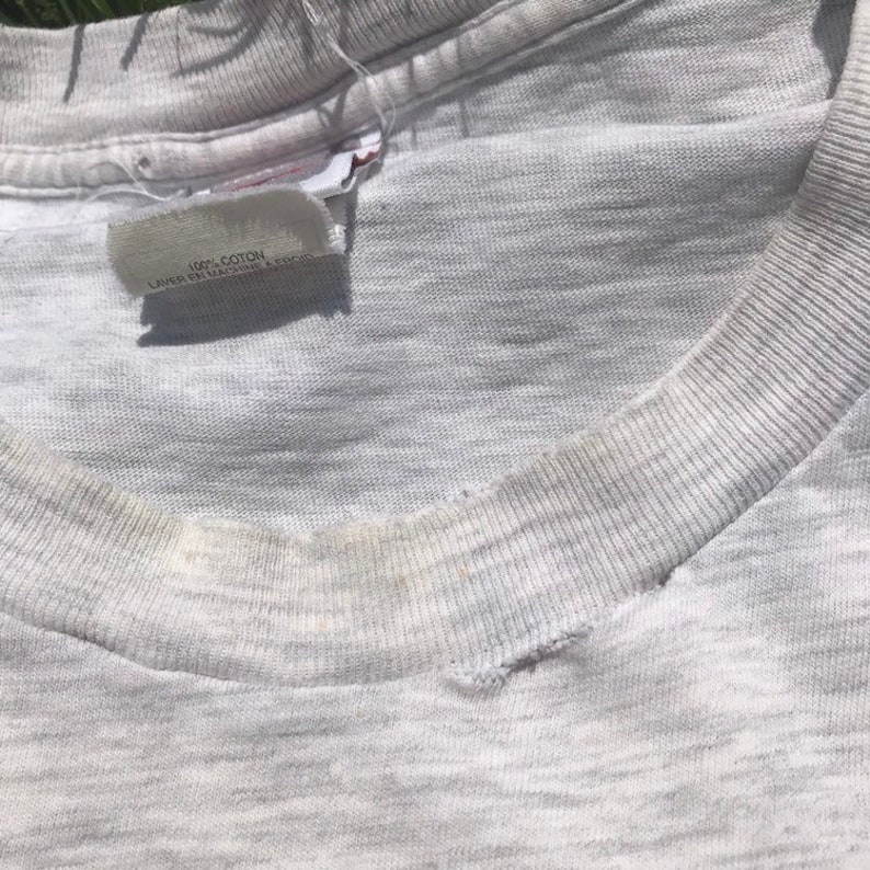 May include: Close-up of a grey t-shirt with a white tag that reads "100% COTTON, LAVEE EN MACHINE A FROID".