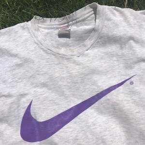 May include: A grey t-shirt with a large purple Nike swoosh logo. The logo is centred on the shirt and has a registered trademark symbol.