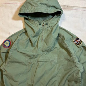 May include: An olive green hooded pullover jacket featuring a high neck with a partial front zipper, a large horizontal zippered chest pocket, and two circular embroidered patches on the sleeves. The right patch depicts a mountain, while the left patch displays the text MANZA.