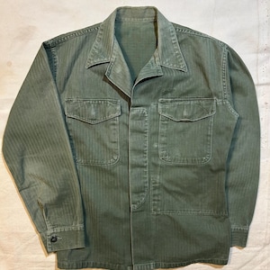 Può includere: Camicia verde oliva in stile militare con due tasche sul petto e chiusura con bottoni. La camicia ha un colletto e maniche lunghe con polsini abbottonati. Il tessuto presenta un sottile motivo a spina di pesce.