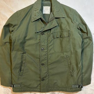 May include: Olive green military-style jacket with a button-up front, two chest pockets, and two side pockets. The jacket has a collared neckline, a zip-up inner lining, and adjustable straps at the waist. The jacket is made of a durable, heavy-duty material.