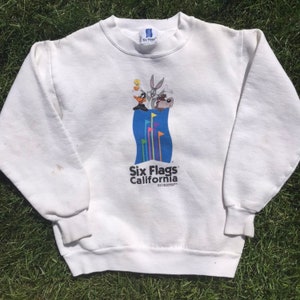 Könnte beinhalten: Weißer Pullover mit einem Vintage-Logo von Six Flags California mit Bugs Bunny, Daffy Duck und Tweety Bird. Das Logo ist blau und zeigt eine bunte Welle mit Flaggen.