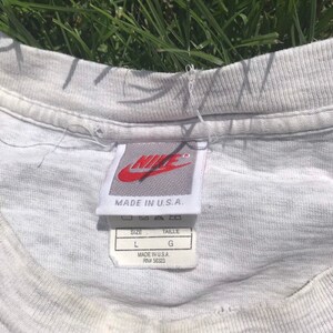 May include: White t-shirt with a grey Nike tag that says "Made in U.S.A." and a size tag that says "Size L, Taille G, Made in U.S.A., RN# 56323"