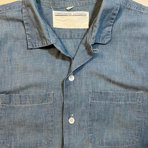 Pode incluir: Uma camisa de cambraia azul-clara com colarinho clássico, fecho frontal com botões brancos e dois bolsos grandes aplicados no peito. Uma etiqueta retangular branca no decote exibe o texto CONSOLIDATED LAUNDRIES.