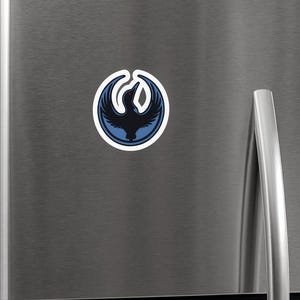 May include: A round sticker featuring a stylized black bird emblem with blue accents, set against a white border. The bird is in a circular design. The sticker is affixed to a stainless steel refrigerator door.