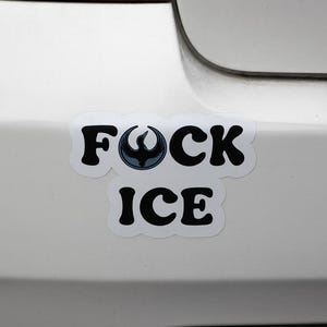 May include: White sticker on a car bumper with the words "FOCK ICE" in black, bold letters. The letter "O" is replaced with a stylized blue and black bird emblem. The sticker is on a white car.