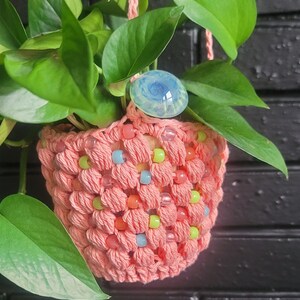 May include: A hanging macrame plant holder in coral pink, adorned with colorful beads. The holder cradles a green leafy plant and is topped with a blue and clear glass decorative piece. The plant hanger is suspended by coral pink cords.