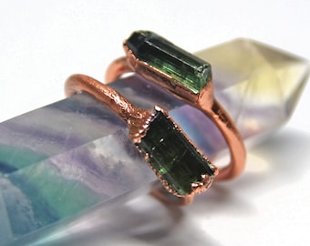 May include: Two copper rings with green tourmaline stones set in the metal. The rings are displayed on a multi-colored fluorite crystal.