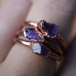 May include: Three copper rings with amethyst gemstones. The rings are stacked on top of each other and are worn on a finger.