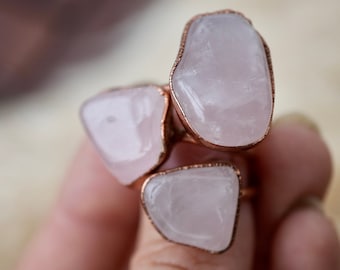 Pode incluir: Três anéis de quartzo rosa com engastes de fio de cobre. Os anéis estão empilhados em um dedo.