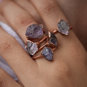 May include: A set of four rings featuring raw amethyst stones wrapped in copper wire. The rings are stacked on a finger.