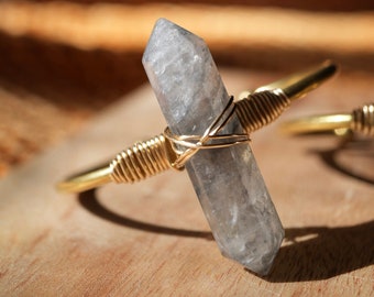 May include: A clear crystal point wrapped in gold wire, creating a unique and delicate bracelet. The crystal has a cloudy, milky appearance and is set against a wooden background.