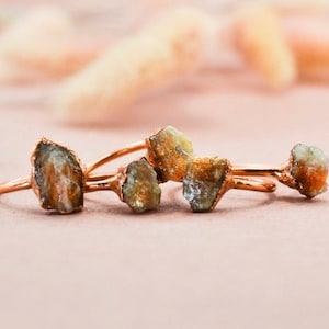 May include: Four rings with raw, natural, brown and green gemstone settings on a copper band. The rings are arranged in a row on a pink surface.