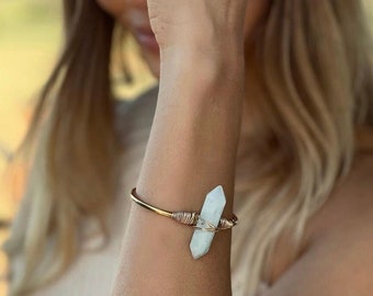 May include: A gold-toned bracelet with a light blue crystal accent. The crystal is wrapped in a wire design. A gold ring is visible on the hand. The person is wearing a beige hat and top.