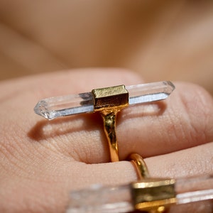 K&ouml;nnte beinhalten: Zwei goldene Ringe mit klaren Quarzkristallspitzen. Die Ringe werden an einem Finger getragen.
