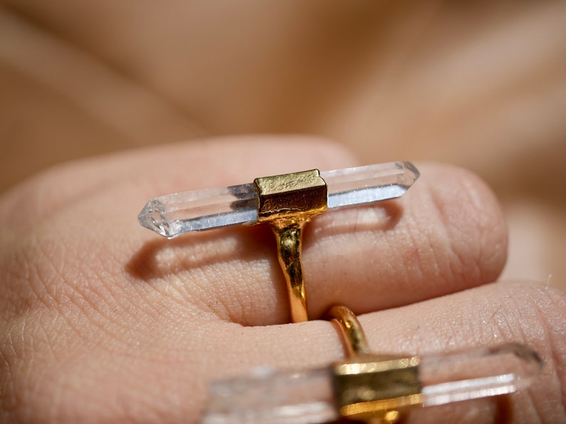 K&ouml;nnte beinhalten: Zwei goldene Ringe mit klaren Quarzkristallspitzen. Die Ringe werden an einem Finger getragen.