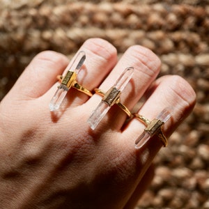 K&ouml;nnte beinhalten: Drei goldene Ringe mit klaren Quarzkristallspitzen. Die Ringe werden an den Fingern einer Hand getragen.