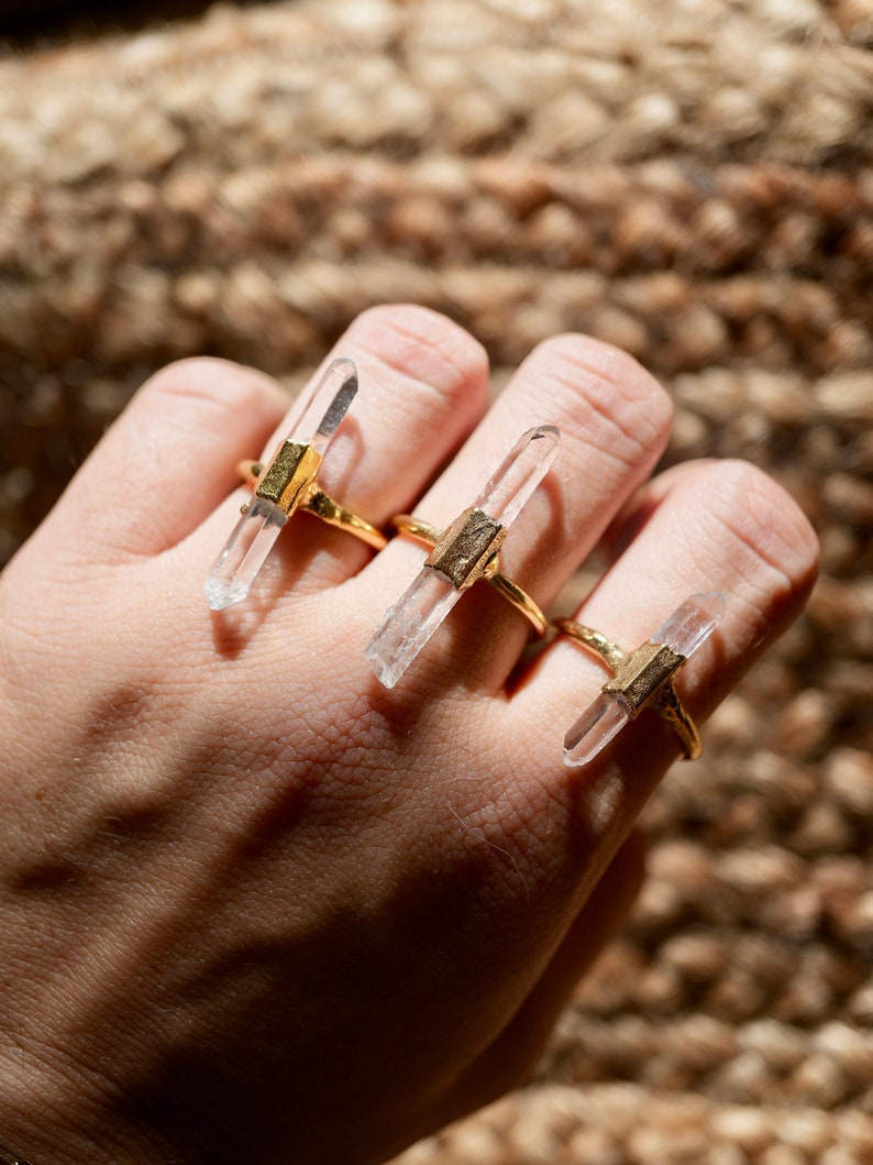 K&ouml;nnte beinhalten: Drei goldene Ringe mit klaren Quarzkristallspitzen. Die Ringe werden an den Fingern einer Hand getragen.