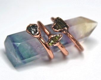 May include: Three copper rings with raw green and black gemstones. The rings are stacked on a green and purple fluorite crystal.