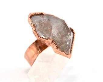 May include: A copper ring with a large, clear quartz stone set in the center. The stone is faceted and has a rough, natural look. The ring is made of hammered copper and has a rustic, handmade feel.