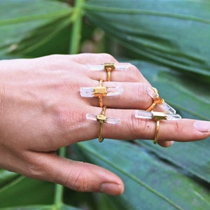 K&ouml;nnte beinhalten: Eine Hand tr&auml;gt f&uuml;nf goldene Ringe mit klaren Quarzkristallsteinen. Die Ringe sind auf den Fingern &uuml;bereinander gelegt. Der Hintergrund ist eine gr&uuml;ne, bl&auml;ttrige Pflanze.