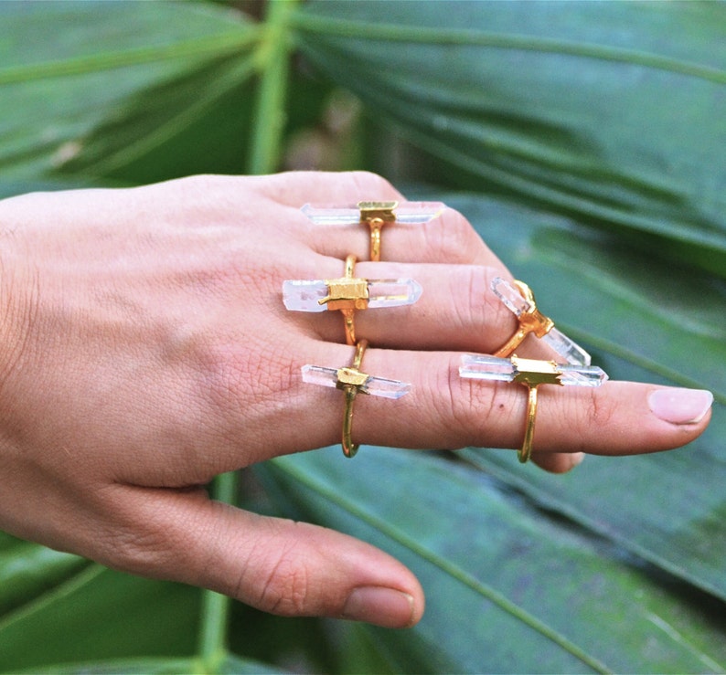 K&ouml;nnte beinhalten: Eine Hand tr&auml;gt f&uuml;nf goldene Ringe mit klaren Quarzkristallsteinen. Die Ringe sind auf den Fingern &uuml;bereinander gelegt. Der Hintergrund ist eine gr&uuml;ne, bl&auml;ttrige Pflanze.