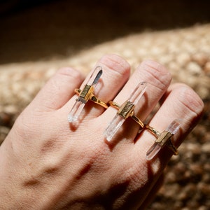 K&ouml;nnte beinhalten: Drei goldene Ringe mit klaren Quarzkristallspitzen. Die Ringe sind auf einem Finger gestapelt.