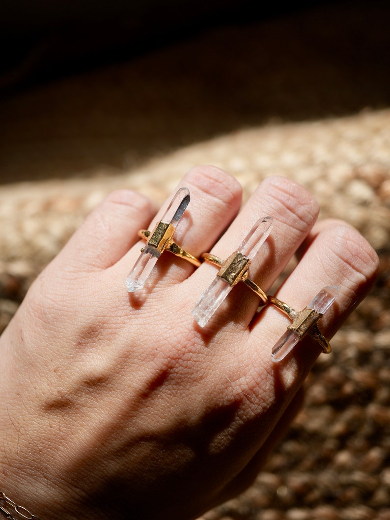 K&ouml;nnte beinhalten: Drei goldene Ringe mit klaren Quarzkristallspitzen. Die Ringe sind auf einem Finger gestapelt.