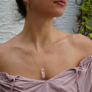 May include: A delicate gold chain necklace with a pale pink crystal pendant. The necklace is worn by a person with a light pink off-the-shoulder top.