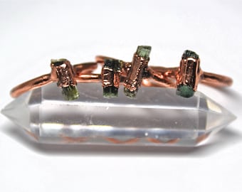 May include: Four copper rings with green and black tourmaline stones set in the metal. The rings are stacked on a clear quartz crystal.
