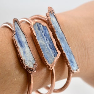 May include: Three copper bracelets with blue kyanite stones. The stones are raw and have a natural, rough texture. The copper is a rose gold color.