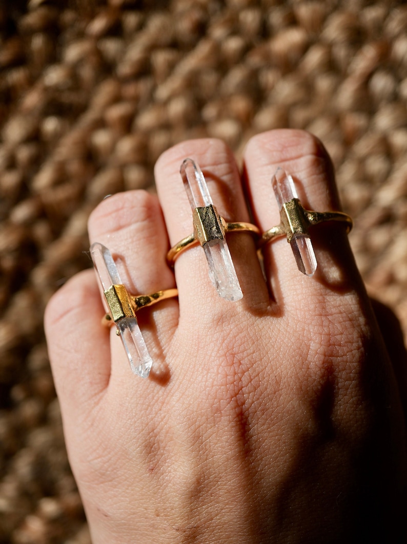 K&ouml;nnte beinhalten: Drei goldene Ringe mit klaren Quarzkristallspitzen. Die Ringe sind auf einem Finger gestapelt.
