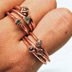 Może przedstawiać: Zestaw czterech miedzianych pierścionków z surowymi akcentami diamentowymi. Pierścionki są ułożone na palcu i mają delikatny i minimalistyczny styl.