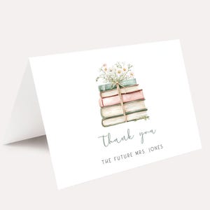 May include: A white folded thank you card with a watercolor illustration of a stack of books tied with a green ribbon and topped with flowers. The text "Thank you" and "THE FUTURE MRS. JONES" are printed in a script font.