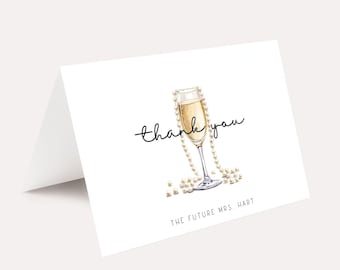 May include: White folded card with a champagne glass illustration. The glass is filled with bubbly liquid and adorned with a pearl necklace. The words "thank you" are written in cursive, with "THE FUTURE MRS. HART" below.