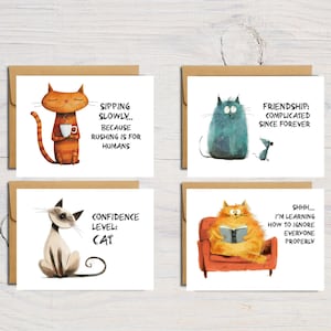 May include: Four greeting cards with cat illustrations. One card shows an orange cat with a coffee cup and the text "Sipping Slowly... Because Rushing is for Humans." Another shows a blue cat with a mouse and the text "Friendship: Complicated Since Forever."