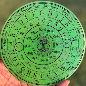 May include: A green circular divination board with a black border and white glitter. The board features the alphabet, numbers, days of the week, months of the year, and the words "YES", "NO", and "POSSIBLY CHECK AGAIN".