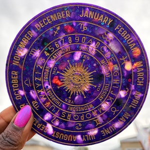 May include: A circular purple resin spirit board featuring gold lettering, including the months of the year, the alphabet, numbers zero through nine, days of the week, and zodiac symbols, with a central sun and eye graphic design.
